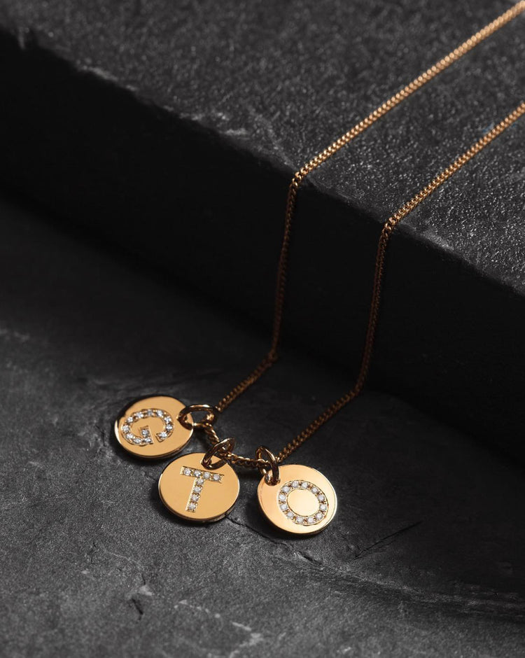 tre guldberlocker med diamant bokstäver i guldkedja placerad på ett svart stenblock. 