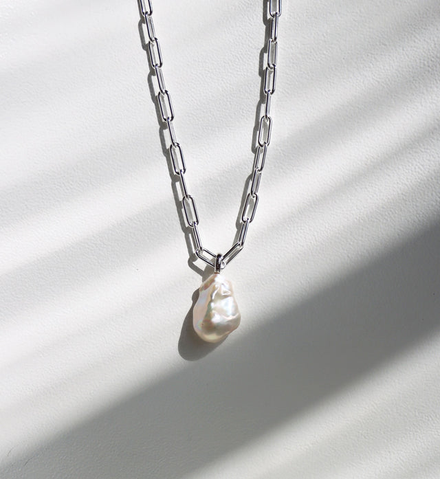 Halsband som hänger mot en solig vägg med en silverkedja som har ovala länkar och en stor oregelbunden barockpärla. 