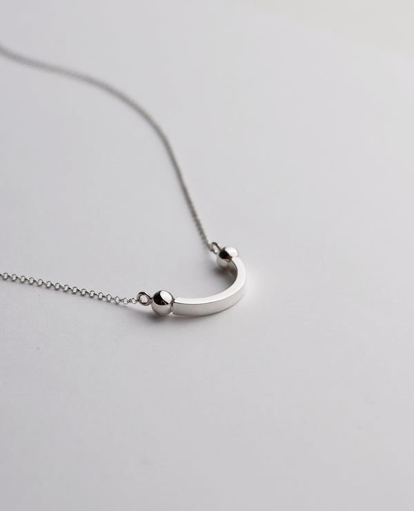Närbild på ett halsband med en båge som har små silverkulor vid ändarna, fäst i en tunn silverkedja. 