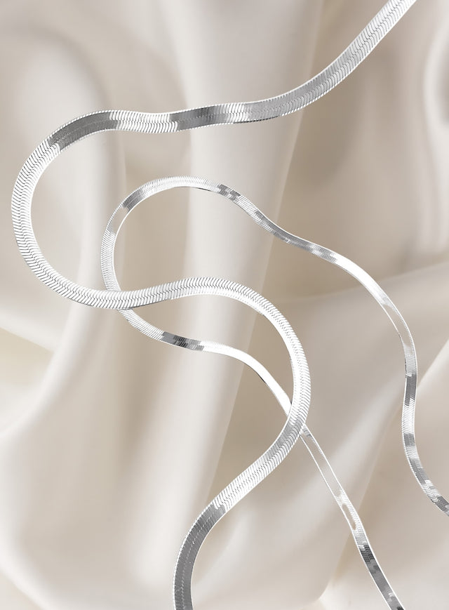 platta silverkedjor av modell snake chain placerade på vitt sidentyg. 