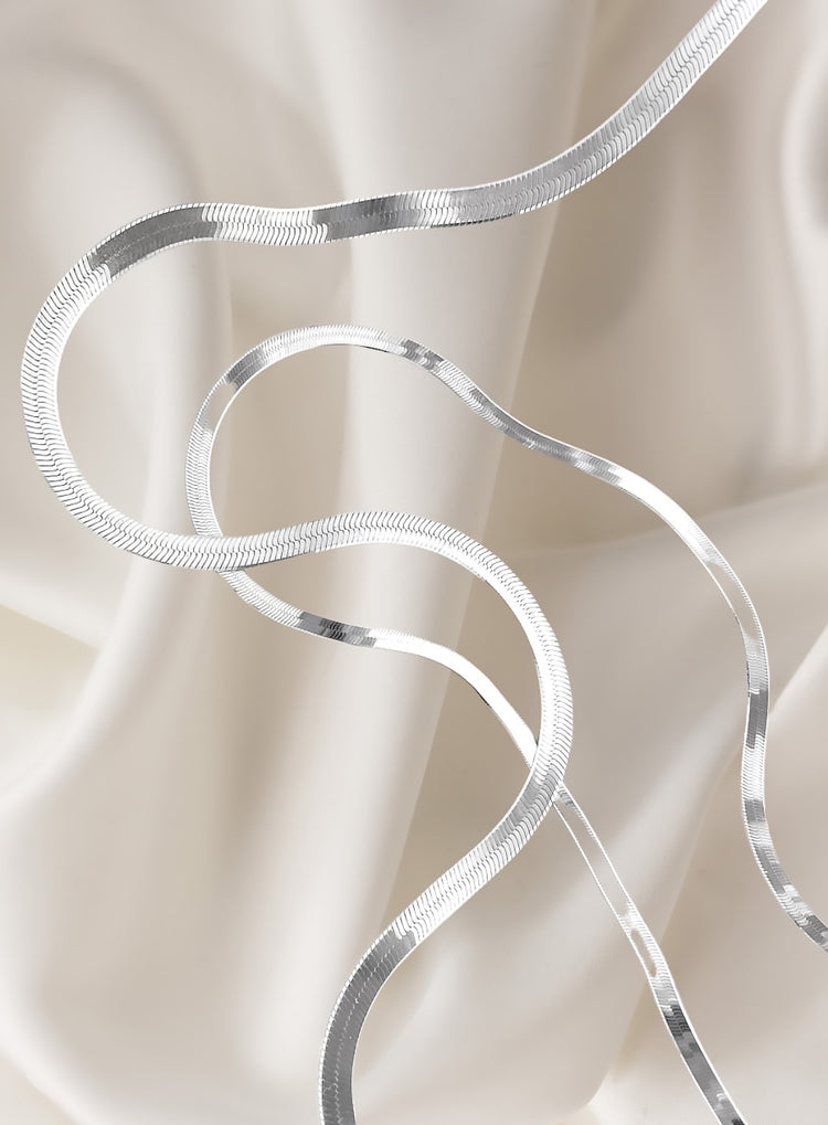 platta silverkedjor av modell snake chain placerade på vitt sidentyg. 