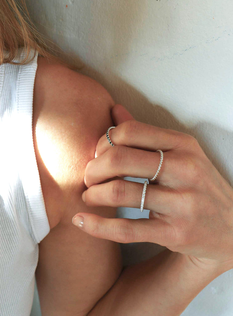Kvinna som bär flera smala ringar på olika fingrar. En smal diamantring med ett band av diamanter, en vriden silverring och en svart diamantring. 