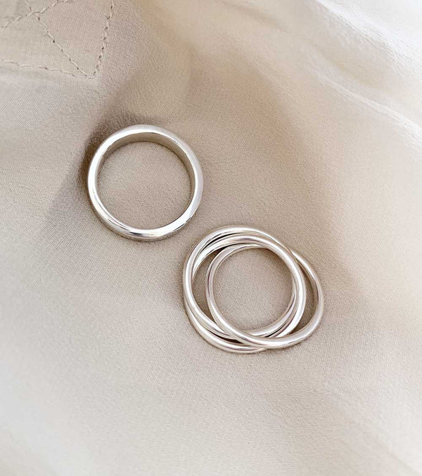 Två silverringar som ligger på ett vitt tyg. En slät silverring och en ring med tre ringar i en ring.